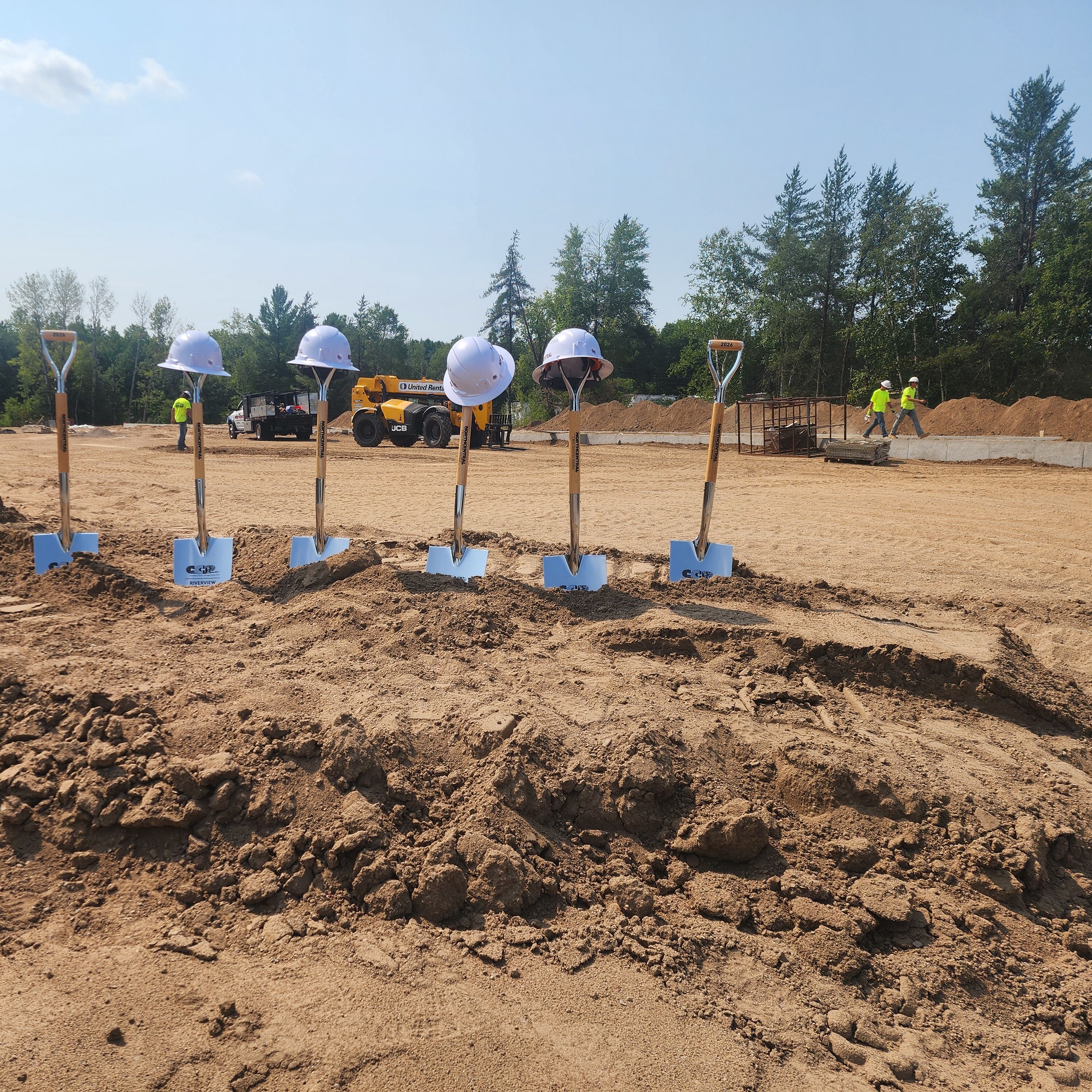 A row of silver shovels and white hard hats lined up on a dirt mound at a construction site, with trees and construction machinery in the background under a bright sky.