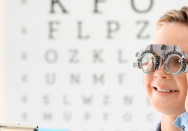 Ein Junge steht vor einer Tafel mit unterschiedlich großen Buchstaben und hat eine Brille vom Augenarzt auf.