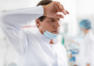 Ein Arzt in weißem Kittel und mit einer chirurgischen Maske unter dem Kinn wischt sich in einem hellen, modernen Krankenhaus oder Labor den Schweiß von der Stirn. Im Hintergrund ist ein weiteres medizinisches Personal in Unschärfe zu sehen.