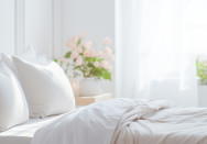 Helles, gemütliches Schlafzimmer mit einem aufgeräumten Bett, weichen weißen Kissen und einer weißen Bettdecke. Im Hintergrund fällt sanftes Tageslicht durch ein großes Fenster herein, das von weißen Vorhängen umrahmt wird.