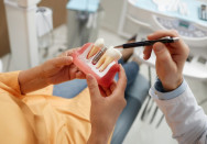 Eine Patientin auf dem Zahnarztstuhl hält ein Teilmodel des Kiefers mit Implantat. Die Hand des Zahnarztes deutet darauf und erklärt.