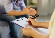 Therapeutin macht Notizen auf einem Klemmbrett während eines Gesprächs mit einem Patienten in einer Beratungssituation zur Burnout-Behandlung und Stressbewältigung in einer ruhigen Praxisumgebung.