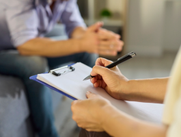 Therapeutin macht Notizen auf einem Klemmbrett während eines Gesprächs mit einem Patienten in einer Beratungssituation zur Burnout-Behandlung und Stressbewältigung in einer ruhigen Praxisumgebung.