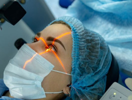 Eine Frau liegt auf einem Behandlungsstuhl und trägt OP-Kleidung. Ihr wird mit einem orangenen Laser in das Auge geleuchtet. 