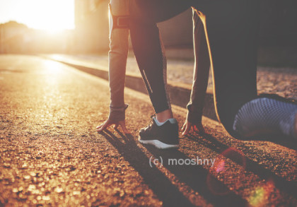 Eine Person trägt Sportkleidung und bereitet sich in einer Startposition auf einem sonnigen, asphaltierten Weg auf einen Lauf vor. Der Sonnenuntergang im Hintergrund wirft lange Schatten und taucht die Szene in ein warmes, goldenes Licht.
