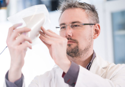 Ein Mann im Labor hält ein Objekt in den Händen und untersucht es sorgfältig. Er trägt ein weißes Laborkittel und eine Brille.