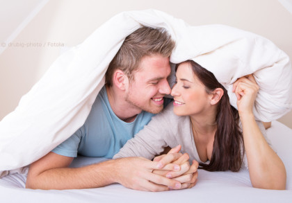 Ein lächelndes Paar liegt auf dem Bett und kuschelt zusammen unter einer weißen Decke. Sie halten sich an den Händen und schauen sich liebevoll an.