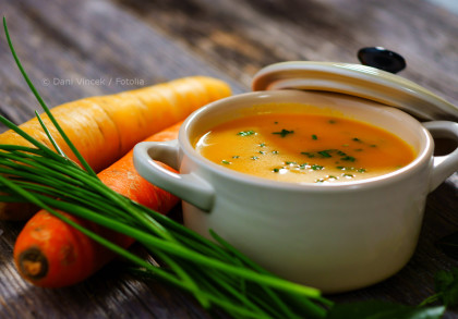 Ein weißer Keramiktopf mit einer gelben Suppe, garniert mit grünen Kräutern, steht auf einem Holztisch. Daneben liegen zwei Karotten und frische Schnittlauchstängel. Der Deckel des Topfes ist leicht geöffnet.