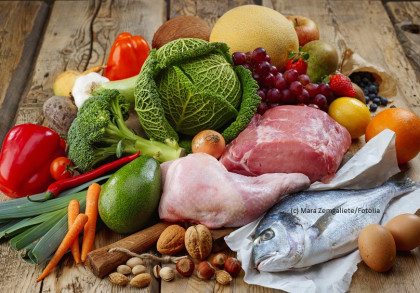 Eine Vielzahl von Lebensmitteln auf einem Holztisch: verschiedene Gemüsesorten wie Paprika, Brokkoli, Karotten, Lauch und Wirsing, eine Avocado, Knoblauch, rote Chilischoten, und Sellerie.