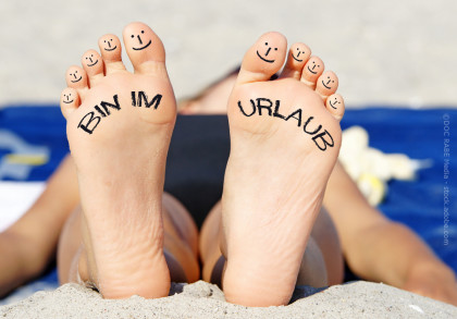 Zu sehen sind die Fußsohlen einer Person am Strand. Auf den Zehen sind lächelnde Gesichter gemalt. Auf der linken Fußsohle steht „BIN IM“ und auf der rechten „URLAUB“, zusammen „BIN IM URLAUB“. Die Person liegt entspannt auf einem blauen Handtuch.