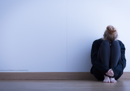 Eine Person sitzt mit angezogenen Knien und verschränkten Armen in einer Ecke eines Raumes auf dem Boden. Die Person trägt dunkle Kleidung und hat das Gesicht auf die Knie gelegt. Der Raum hat weiße Wände und einen Holzboden.