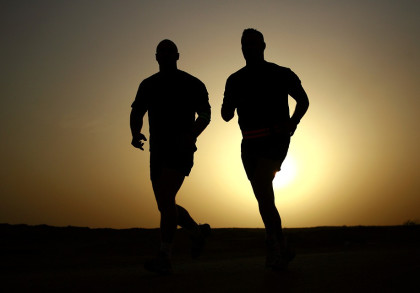 Zwei Personen joggen bei Sonnenuntergang. Ihre Silhouetten sind vor der untergehenden Sonne sichtbar. Der Himmel ist düster mit einem Hauch von Orange und Gelb.