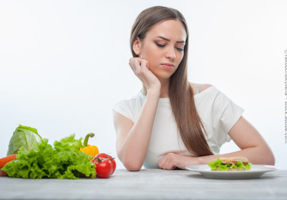 Eine Frau mit langen braunen Haaren sitzt an einem Tisch und stützt ihren Kopf nachdenklich auf ihre Hand. Vor ihr auf dem Tisch liegen verschiedene Gemüsesorten wie ein Kopfsalat, Tomaten, eine Karotte, ein Kohl und eine Paprika.