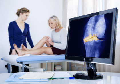 Ein Arzt untersucht das Knie einer Patientin, die auf einem Untersuchungstisch sitzt. Im Vordergrund auf einem Schreibtisch steht ein Monitor, auf dem eine Röntgenaufnahme eines Knies zu sehen ist. Daneben liegen einige Dokumente und ein Kugelschreiber.