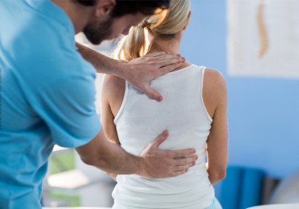 Ein Physiotherapeut behandelt den oberen Rücken einer Frau in einem Behandlungsraum. Die Frau trägt ein weißes Tanktop und sitzt mit dem Rücken zur Kamera, während der Therapeut in einem blauen Hemd seine Hände auf ihren Rücken legt.