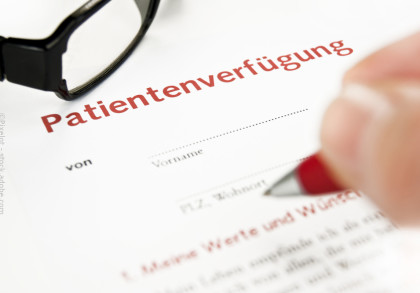 Nahaufnahme einer Hand, die dabei ist, mit einem Stift ein Dokument mit dem Titel "Patientenverfügung" auszufüllen. Eine Brille liegt neben dem Dokument.