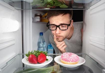 Ein Mann mit Brille schaut nachdenklich in einen Kühlschrank. Im Vordergrund befinden sich auf einem Regal zwei Teller, einer mit frischen Erdbeeren & einer mit einem rosa glasierten Donut. Im Hintergrund stehen eine Wasserflasche und andere Lebensmittel.