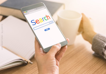Eine Person hält ein Smartphone in der Hand, das eine Suchmaschine mit dem Wort "Search" auf dem Bildschirm anzeigt. Im Hintergrund sind ein geöffnetes Notizbuch und eine Tasse Kaffee zu sehen.