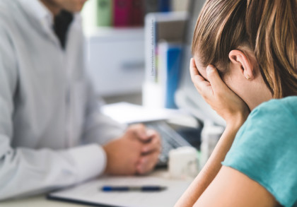 Eine Frau sitzt mit dem Gesicht in den Händen und scheint verzweifelt oder traurig zu sein. Vor ihr sitzt eine Person, wahrscheinlich ein Arzt oder Berater, der seine Hände gefaltet vor sich auf dem Tisch liegen hat.