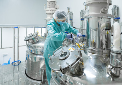 Eine Person in Schutzkleidung, einschließlich Haarnetz, Maske und Handschuhen, arbeitet in einer modernen Produktionsanlage mit glänzenden Edelstahlbehältern und Maschinen.