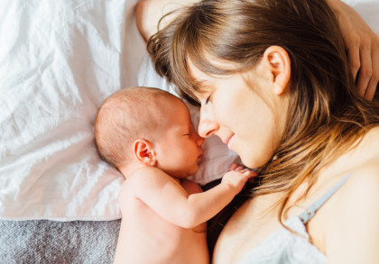 Eine Frau liegt auf einem Bett und hält ein neugeborenes Baby eng an ihrem Gesicht. Beide haben die Augen geschlossen und sehen friedlich aus. Die Frau hat lange braune Haare, und das Baby ist nackt. Sie liegen auf weißen Laken und einer grauen Decke.