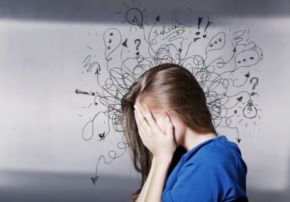 Eine Frau hält sich den Kopf in den Händen und wirkt gestresst. Hinter ihr sind viele chaotische Kritzeleien, wie Glühbirnen, Ausrufezeichen und Fragezeichen, gezeichnet, die ihren unruhigen Geisteszustand symbolisieren. Sie trägt ein blaues Oberteil.