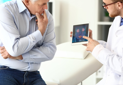 Ein älterer Mann sitzt in einer Arztpraxis und scheint nachdenklich, während ein Arzt auf ein Tablet zeigt, das eine medizinische Grafik der Harnwege anzeigen. Der Arzt trägt einen weißen Kittel und eine Brille.