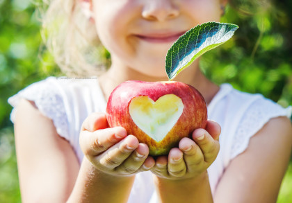 Ein Kind hält einen roten Apfel in beiden Händen. Der Apfel hat einen herzförmigen Biss in der Mitte. Der Hintergrund ist verschwommen und zeigt grüne Blätter und Bäume. Das Kind trägt ein weißes Oberteil und lächelt.