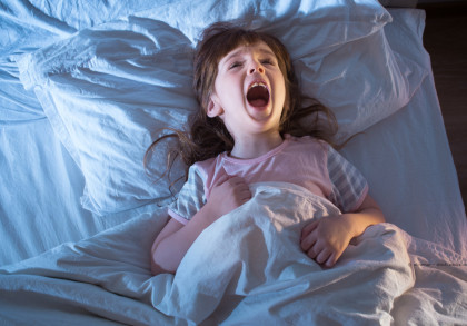 Ein kleines Mädchen liegt in einem Bett und scheint erschrocken oder verängstigt zu sein. Sie hat den Mund weit geöffnet und schreit. Sie trägt ein rosa Schlafanzug-Oberteil und hält die Bettdecke fest. Die Bettwäsche und das Kopfkissen sind weiß.