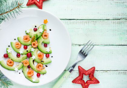 Eine kreative Darstellung eines Weihnachtsbaums auf einem Teller, gemacht aus Avocadoscheiben, Lachsstückchen, Sternförmigen Käsestücken, roten Johannisbeeren und Pfefferkörnern.