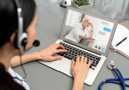 Eine Person mit einem Headset führt eine Telemedizin-Beratung über ihren Laptop durch. Auf dem Laptop-Bildschirm ist eine ältere Frau zu sehen, die anscheinend Kopfschmerzen hat und ihr Kopf mit ihrer Hand stützt.