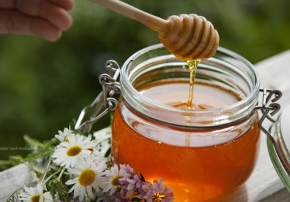 Eine Hand hält eine Honigwabe über ein offenes Glas mit goldenem Honig, aus dem Honig tropft. Neben dem Glas liegen einige weiße Gänseblümchen. Die Szene ist draußen und im Hintergrund ist grüne Vegetation zu sehen.