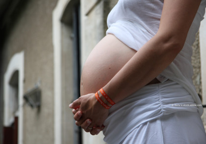 Eine schwangere Frau, die ein weißes Shirt und eine weiße Hose trägt und von der Seite fotografiert wird. Sie hält ihre Hände vor ihrem Bauch zusammengefaltet. Im Hintergund ist ein unscharfes Gebäude zu sehen.