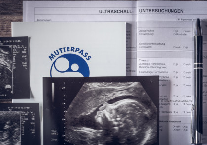 Ein Bild zeigt mehrere Ultraschallbilder und ein ärztliches Dokument. Im Vordergrund liegt ein Mutterpass mit einem blauen Symbol darauf. Ein Kugelschreiber befindet sich auf dem Dokument mit der Überschrift "Ultraschall-Untersuchungen".
