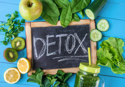 Tafel mit der Aufschrift "DETOX" umgeben von frischem Obst und Gemüse, darunter ein grüner Apfel, Spinatblätter, Gurkenscheiben, Blattsalat, Kiwischeiben, Zitronenscheiben und ein Smoothie in einem Glas mit gelbem Deckel und Strohhalm.