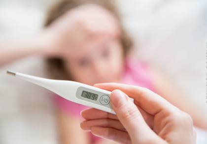 Eine Hand hält ein digitales Thermometer, das eine Temperatur von 38,9 °C anzeigt. Im Hintergrund liegt ein Kind mit einer Hand auf der Stirn und sieht krank aus.