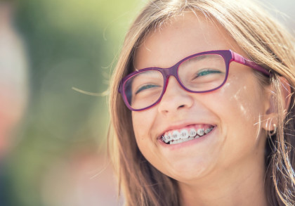 Ein lächelndes Mädchen mit einer Zahnspange und lila Brille. Hintergrund unscharf mit grünlichen Tönen. Gesichtsnahaufnahme bei Tageslicht.