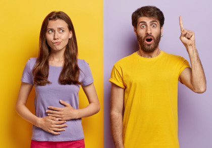 Ein junges Paar steht vor einer zweifarbigen Wand, die in Gelb und Lila geteilt ist. Die Frau links trägt ein lila T-Shirt und fasst sich mit besorgtem, überlegendem Gesichtsausdruck an den Bauch.