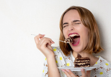 Eine Frau mit schulterlangem braunem Haar hält in der linken Hand eine kleine weiße Untertasse mit einem Stück Schokoladentorte, in der rechten eine Gabel mit Torte, das sie gleich genießen will. Die Frau trägt ein weißes Oberteil mit gelbem Blumenmuster.