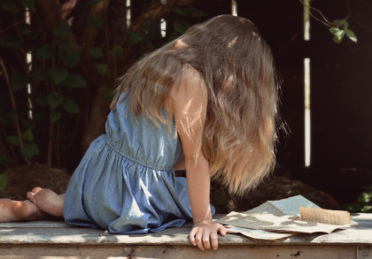 Ein kleines Mädchen mit langen, welligen Haaren sitzt auf einem Holztisch und liest Papiere. Sie trägt ein blaues Kleid und ist in einem schattigen Garten mit Bäumen und Pflanzen umgeben. Die Atmosphäre wirkt ruhig und friedlich.
