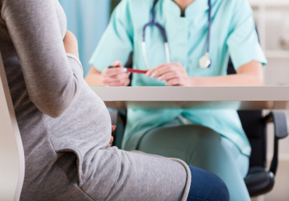 Schwangere Frau in grauem Oberteil sitzt vor einem Mediziner in türkisfarbener Arbeitskleidung mit Stethoskop um den Hals und hält einen Stift in der Hand, während sie eine Unterhaltung führen.