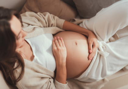Eine schwangere Frau liegt auf dem Sofa und hält sanft ihren nackten Babybauch mit ihren Händen. Sie trägt bequeme, helle Kleidung.