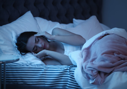 Eine Frau liegt im Bett und hält sich die Hände an den Kopf, als hätte sie Kopfschmerzen. Sie scheint unter einer Decke zu liegen, und der Raum ist dunkelblau beleuchtet.
