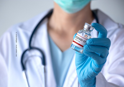 Eine Ärztin hält ein Fläschchen mit COVID-19-Impfstoff in einer behandschuhten Hand. Im Hintergrund ist der Oberkörper der Ärztin  in einem weißen Kittel mit Stethoskop und Maske sichtbar.