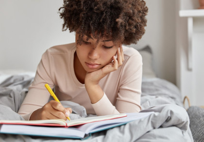 Eine junge Frau liegt auf dem Bett und schreibt in ein Notizbuch. Sie trägt ein weißes Oberteil und hält einen gelben Stift in der Hand. Ihr Gesichtsausdruck ist konzentriert, und sie stützt ihren Kopf mit einer Hand ab. Der Hintergrund ist unscharf.