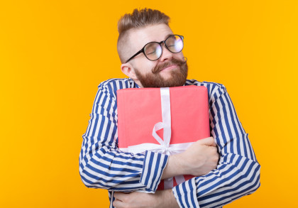 Ein Mann mit Brille und gestreiftem Hemd umarmt lächelnd ein großes, rotes Geschenk mit weißer Schleife vor einem orangefarbenen Hintergrund.