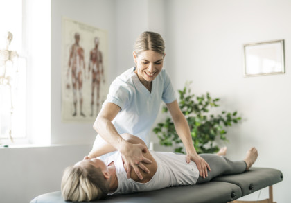 Eine Physiotherapeutin behandelt eine liegende Patientin auf einer Therapieliege. Die Therapeutin lächelt, während sie an der Schulter ihrer Patientin arbeitet. Im Hintergrund sind ein anatomisches Poster und eine Pflanze zu sehen.