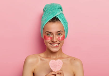 Eine lächelnde Frau mit einer türkisfarbenen Handtuch-Turban auf dem Kopf hat rosa Augenpads unter ihren Augen und hält einen herzförmigen Schwamm. Sie steht vor einem rosa Hintergrund.