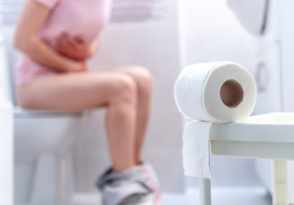 Eine Person sitzt auf einer Toilette und hält sich den Bauch. Es gibt eine nahe Ansicht einer Toilettenpapierrolle im Vordergrund. Die Hose der Person ist heruntergelassen und liegt um ihre Knöchel.