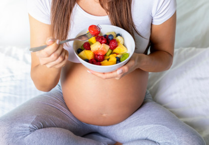 Eine schwangere Frau sitzt auf einem Bett und hält eine Schüssel mit frischem Obstsalat in der Hand. Sie isst mit einer Gabel ein Stück Obst. Sie trägt ein weißes T-Shirt und graue Hosen.